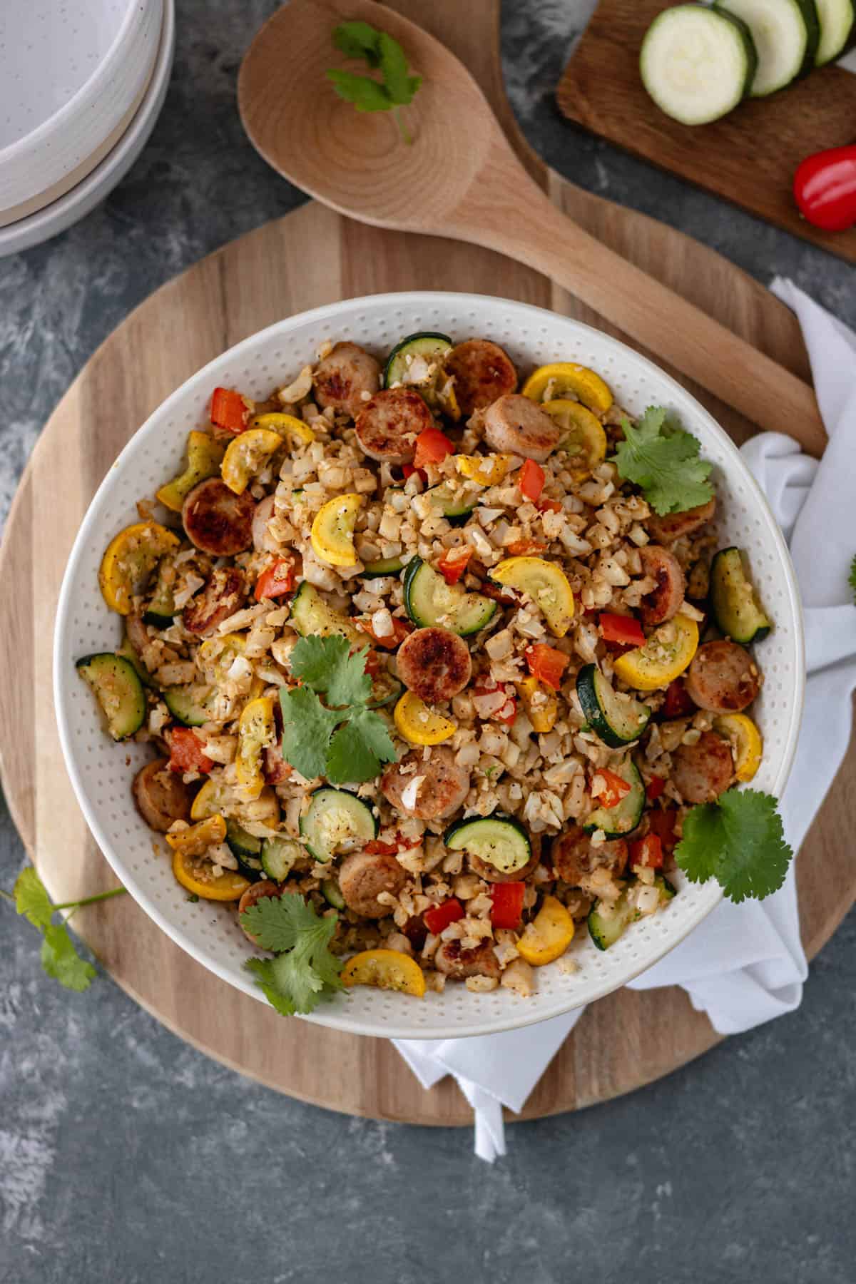 A ceramic serving bowl filled with cooked cauliflower rice, chicken sausage, sliced zucchini, squash, bell pepper and topped with fresh cilantro sprigs, that is sitting on a circular bread board, and surrounded by a white tea towel, serving spoon, ceramic bowls and a mini cutting board with mini bell peppers and sliced zucchini on top.