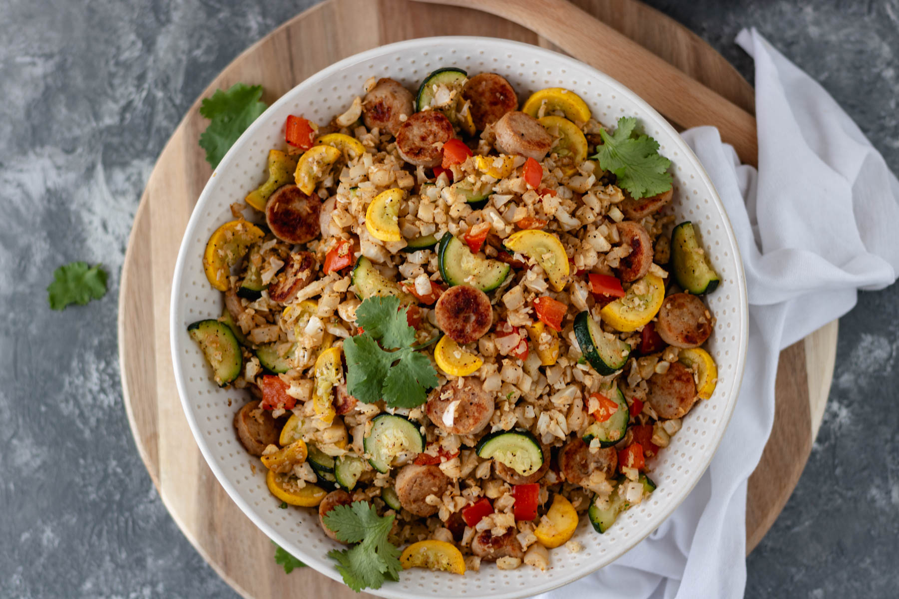 A large ceramic serving bowl filled with cooked cauliflower rice, chicken sausage, sliced zucchini, squash, bell pepper and topped with fresh cilantro sprigs, that is sitting on a circular bread board with a tea towel.