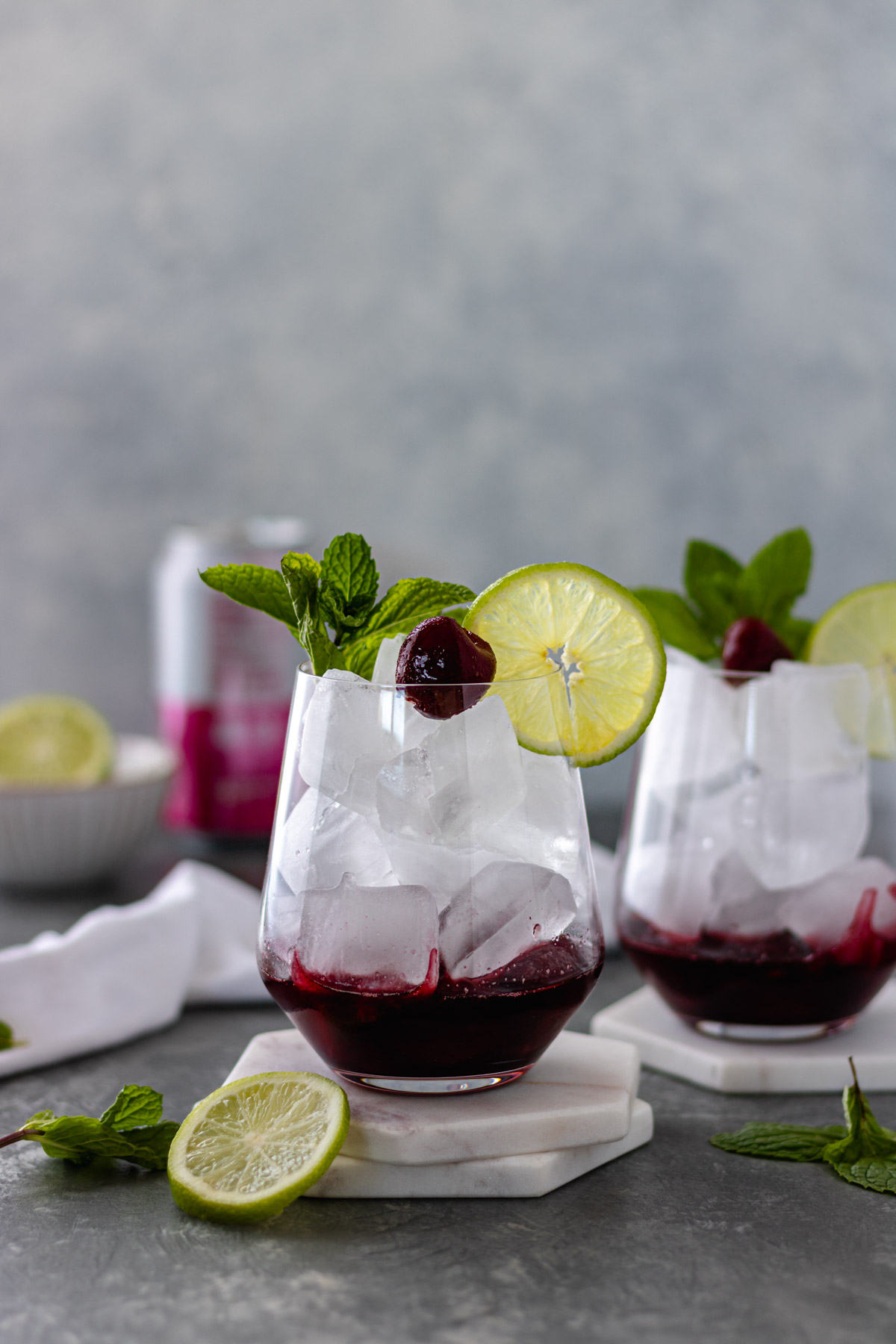 Two glasses of ice and cherry juice topped with a frozen cherry, lime slice and a sprig of mint, surrounded by fresh mint sprigs, a lime slice, white tea towel, a pinch bowl with a lime and a can of sparkling water.