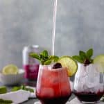 A hand pouring a can of sparkling water into a glass of cherry juice sitting on top of two ceramic coasters, surrounded by another glass of cherry juice, mint sprigs, lime slices, a pinch bowl with a lime and a can of sparkling water.