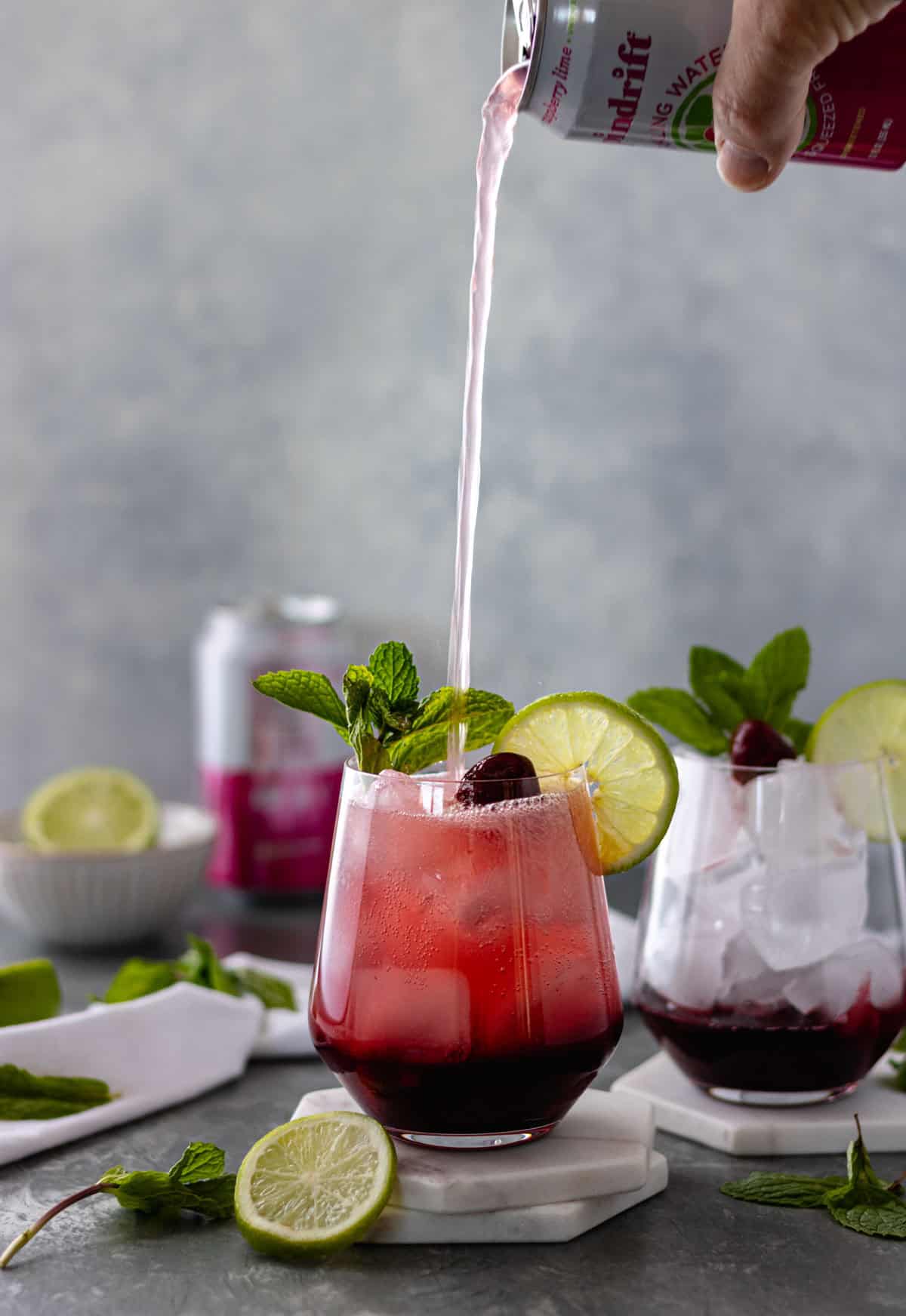 A hand pouring a can of sparkling water into a glass of cherry juice sitting on top of two ceramic coasters, surrounded by another glass of cherry juice, mint sprigs, lime slices, a pinch bowl with a lime and a can of sparkling water.