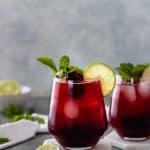 Two full glasses of a Sparkling Cherry Lime Mocktail topped with cherries, a lime slice, and a sprig of mint, sitting on ceramic coasters, and surrounded by mint sprigs, a white tea towel and a pinch bowl with a lime.