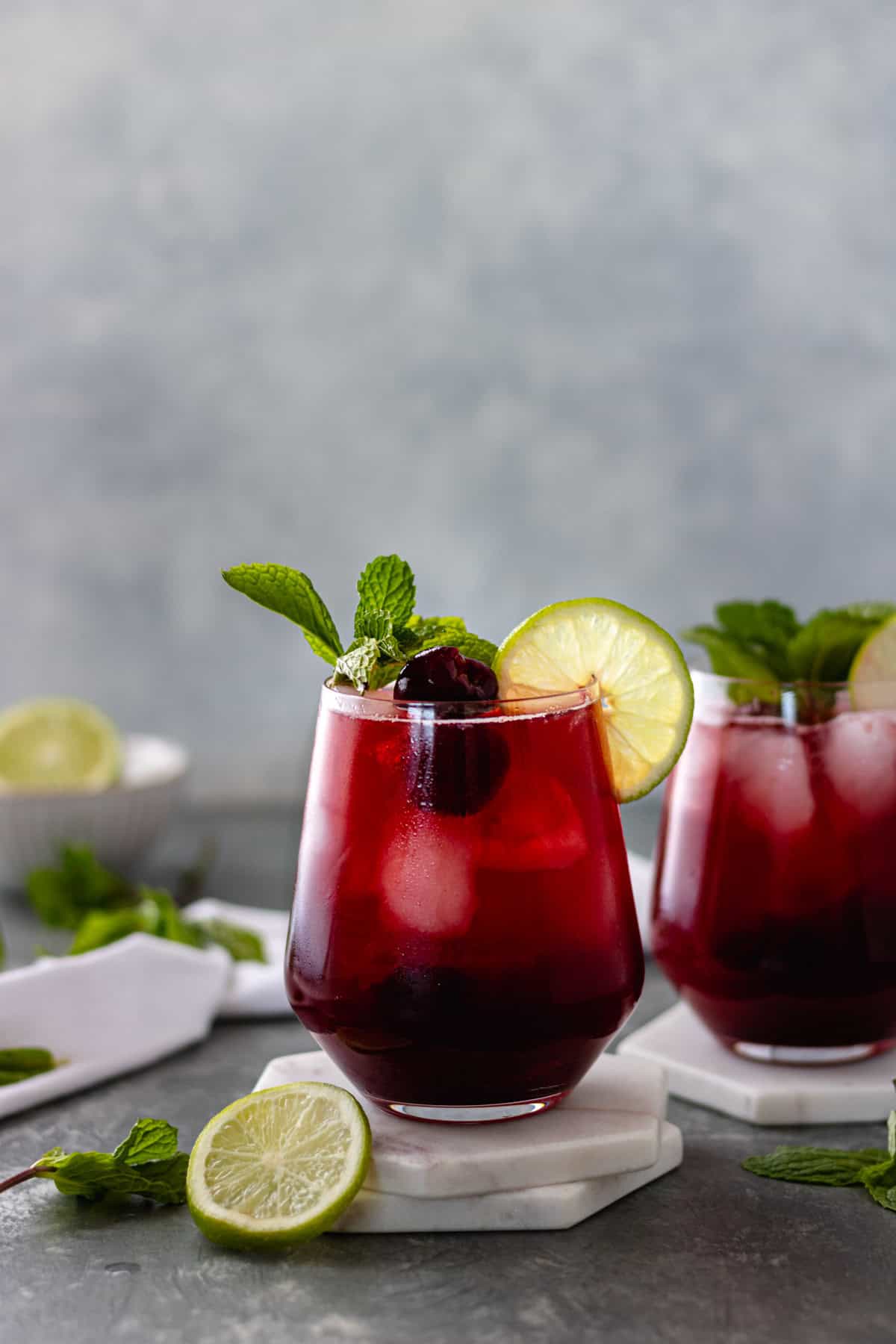 Two full glasses of a Sparkling Cherry Lime Mocktail topped with cherries, a lime slice, and a sprig of mint, sitting on ceramic coasters, and surrounded by mint sprigs, a white tea towel and a pinch bowl with a lime.